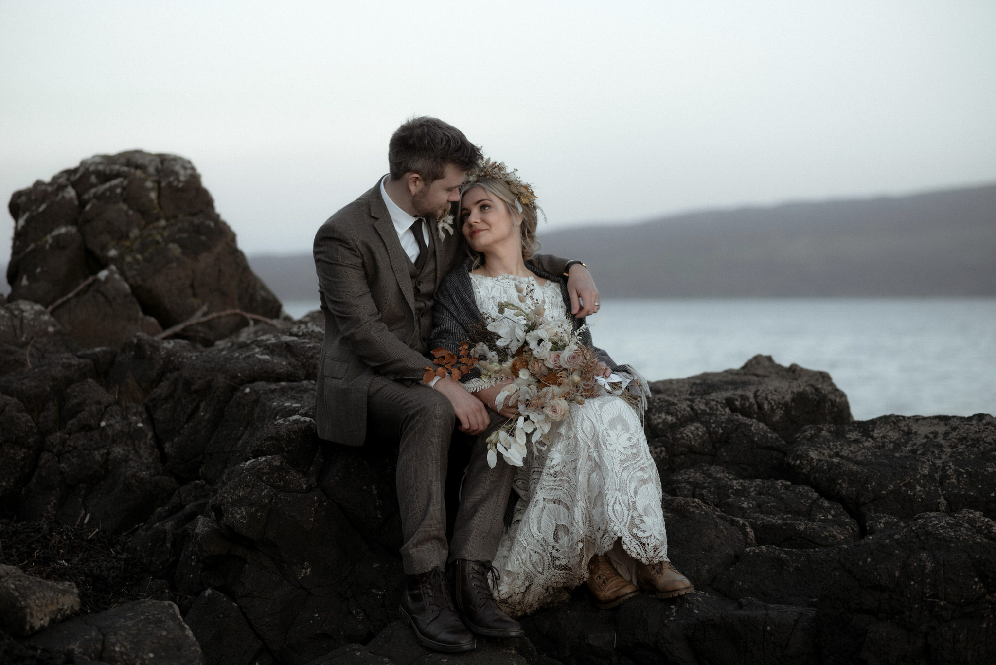 An eloping couple sit on black rocks arm in arm by the sea on the Isle of Skye at blue hour on their elopement day