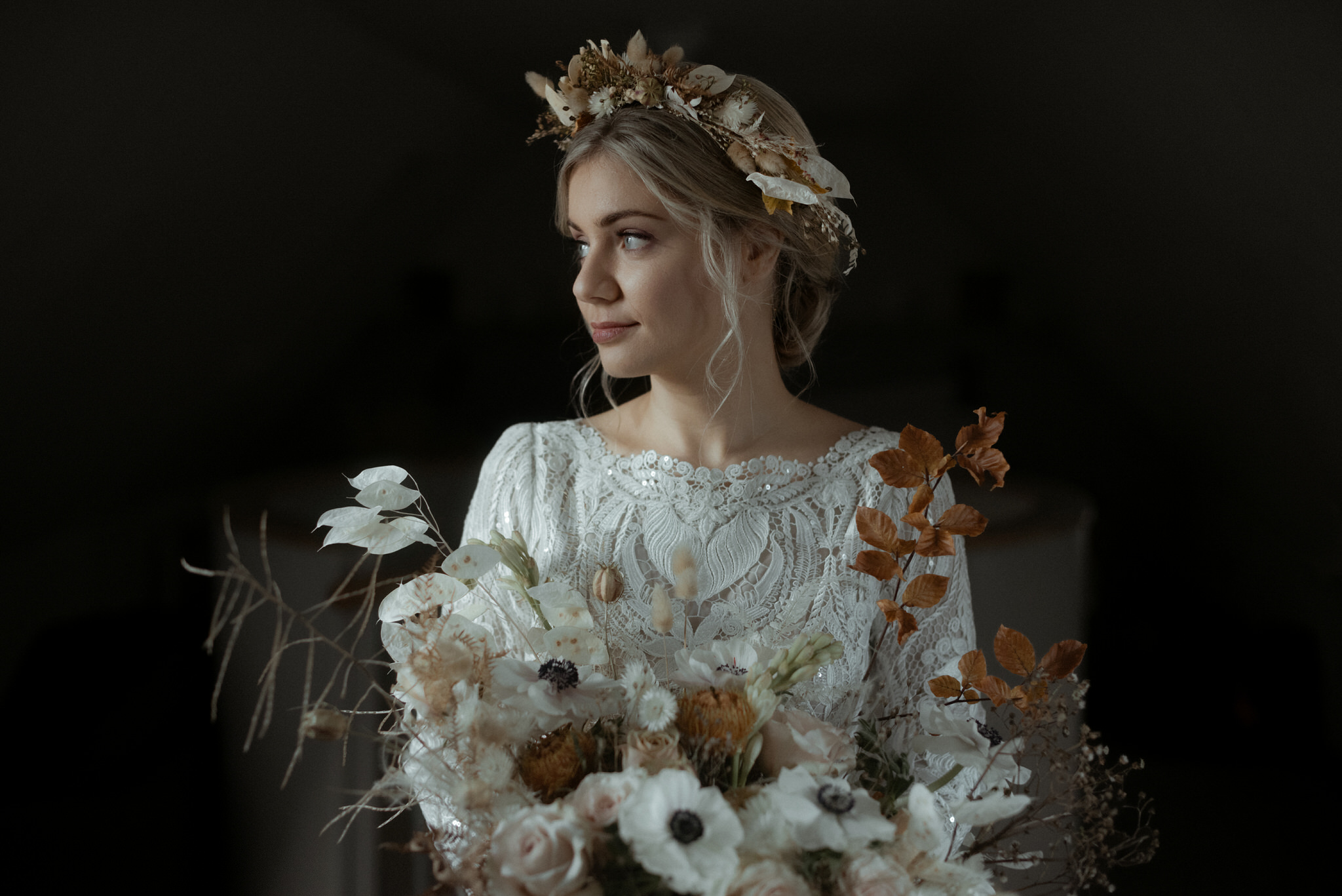 A portrait of a bride stood in a dark room holding flowers of honesty birch leaves and roses looks to the left of the frame into the available light